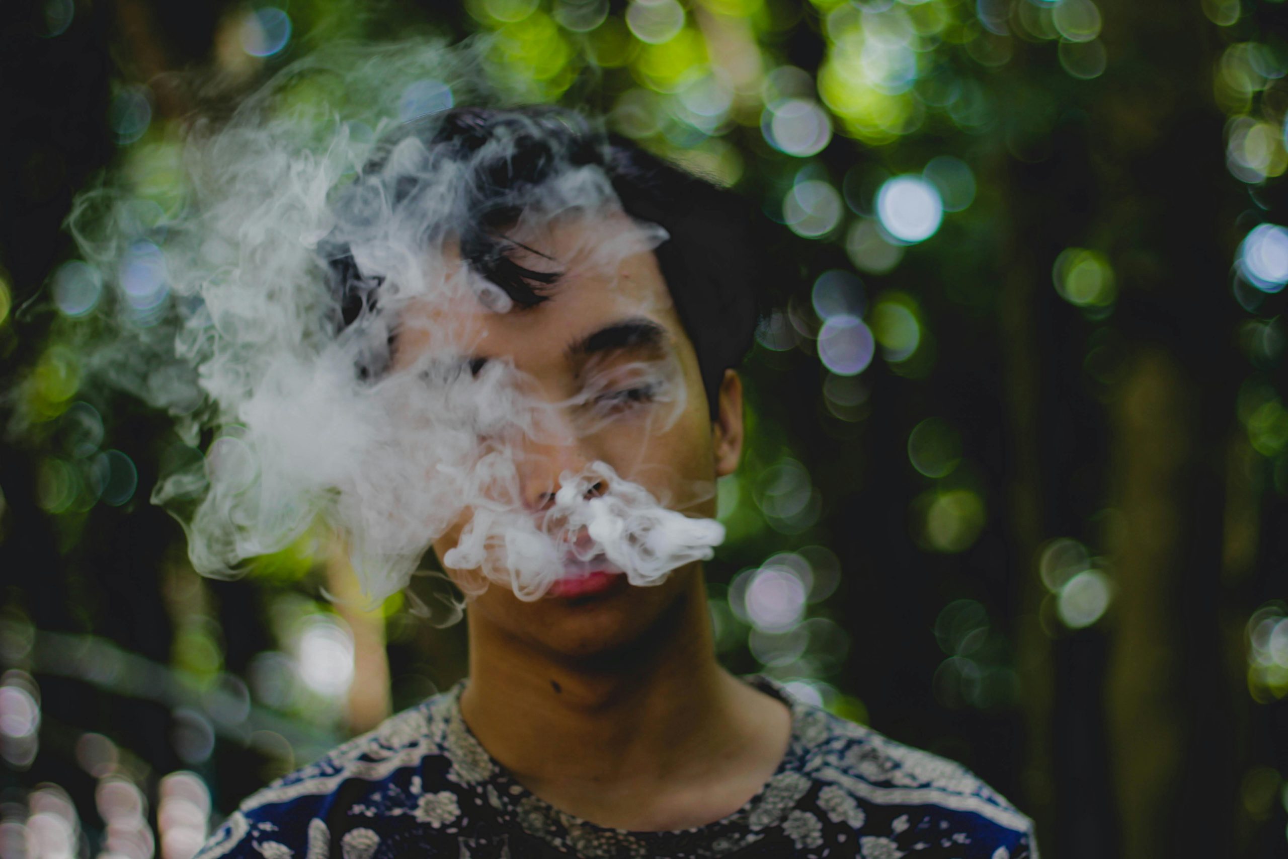 A young man smoking and exhaling smoke outdoors with a blurred natural background.